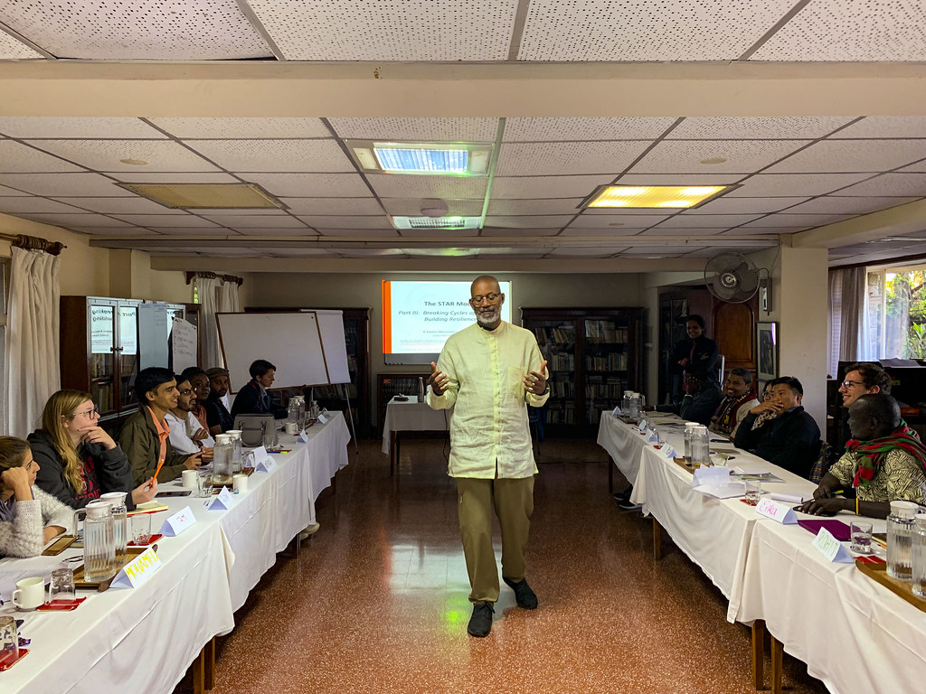 David facilitating a gathering between two long tables, surrounded by participants