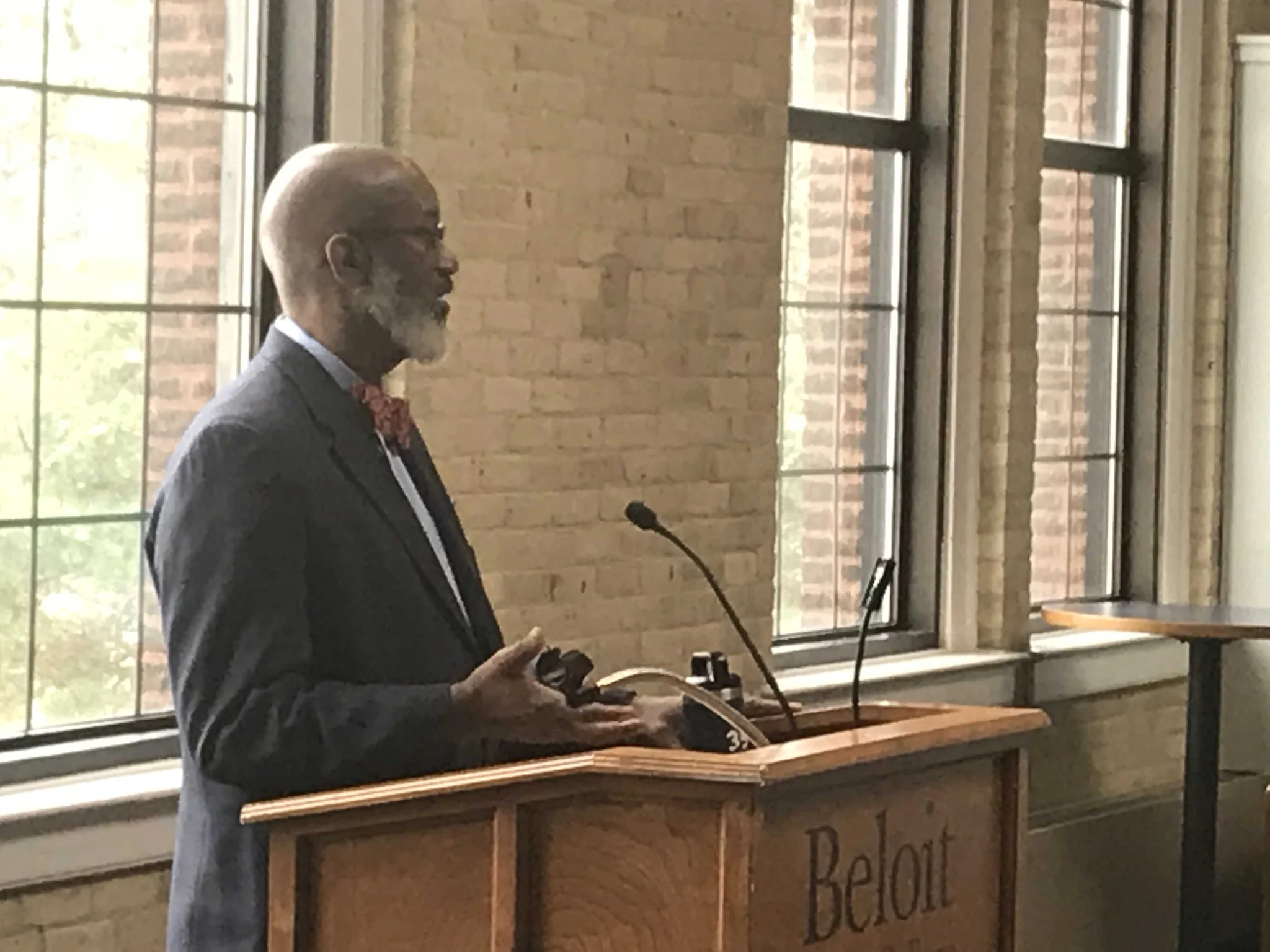 David speaking at a wooden lectern beside tall windows and a brick wall.